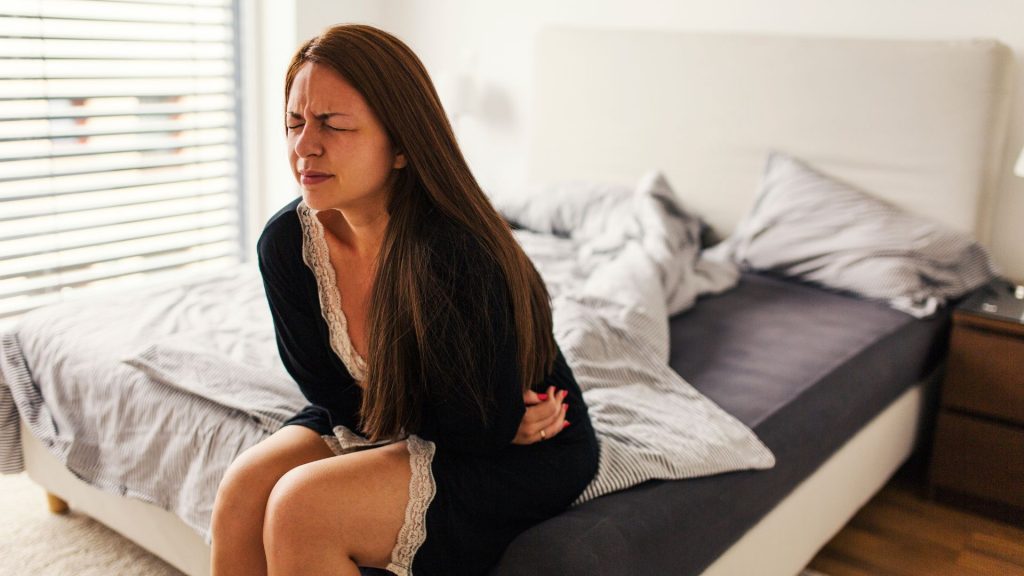 Kobieta siedzi na łóżku i łapie się za brzuch; prawdopodobnie odczuwa ból jajników po stosunku.